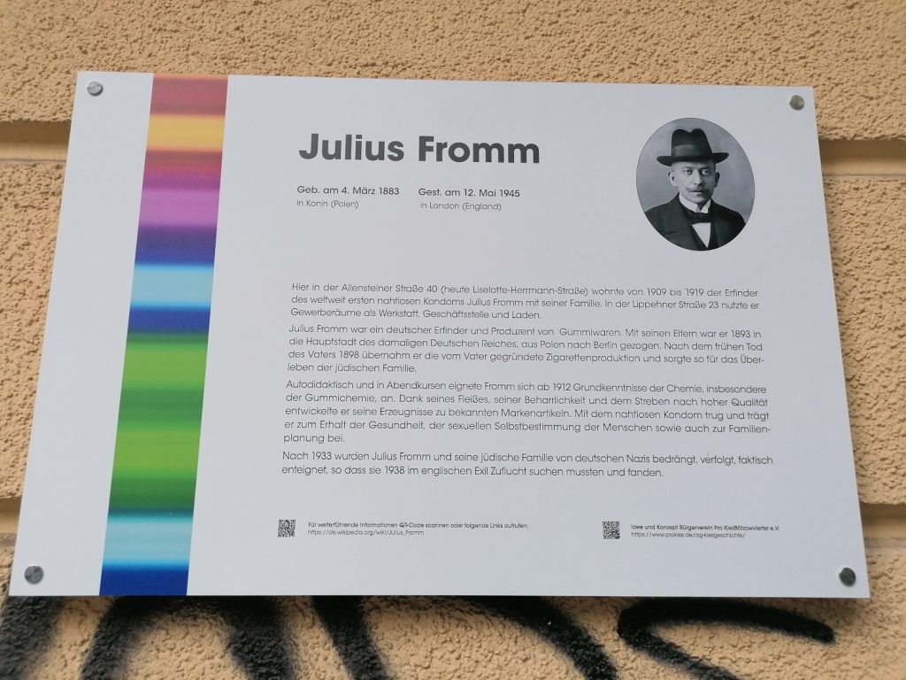 Tafel mit Foto, buntem Streifen und Text zu Julius Fromm an Hauswand